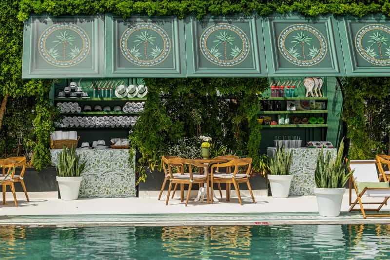 Pavillon bar vert sauge au soleil, panneaux à palmiers, murs de lierre et chaises en bois derrière une piscine turquoise