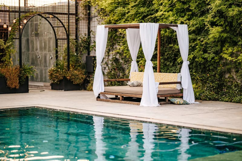 Un lit de repos en bois à rideaux blancs au bord d'une piscine turquoise, devant un mur de lierre et une arche métallique éclairée