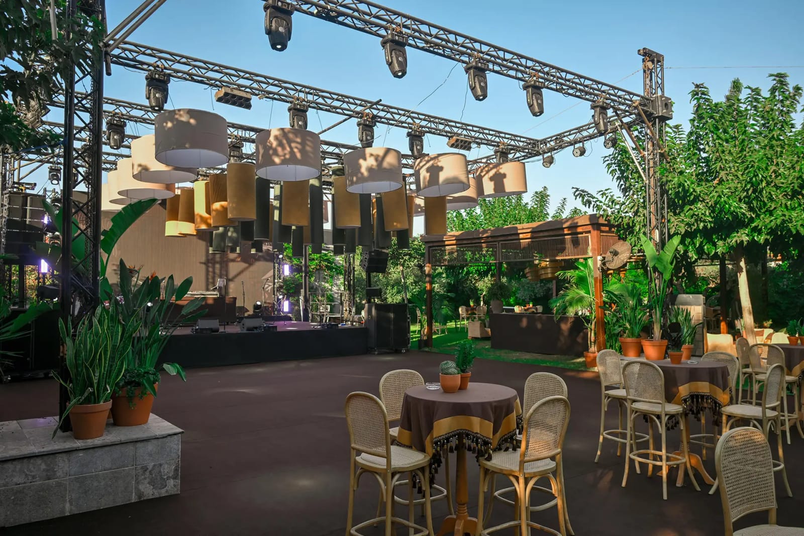 Scène en plein air avec structure d'éclairage et abat-jours suspendus, tables de cocktail aux chaises en rotin et arbres fruitiers