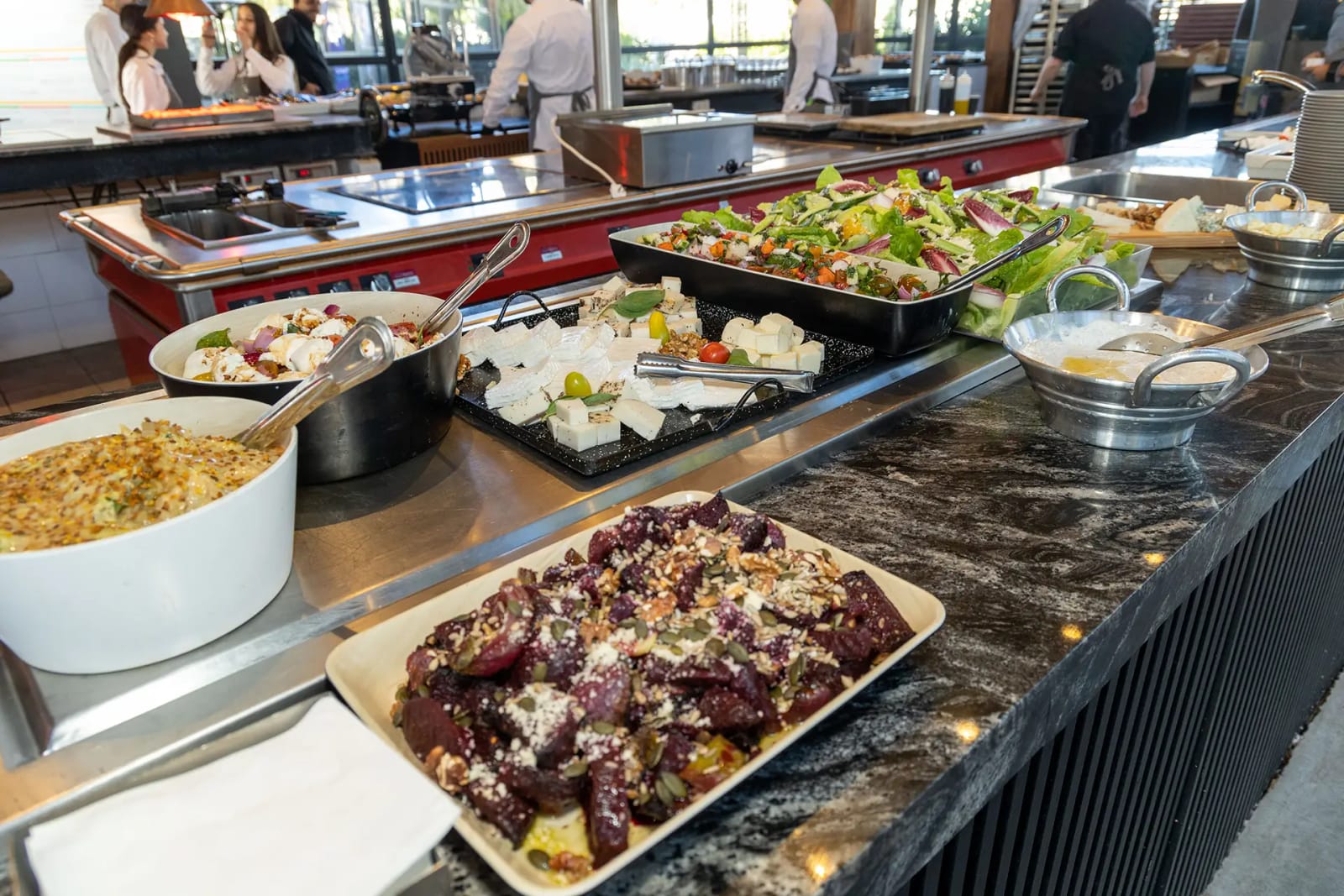 Hot and cold buffet dishes on a black marble counter, with chefs working at an open kitchen in the background