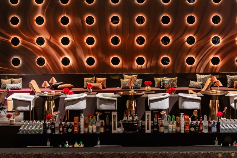 Long lounge counter with white armchairs, bottles and red flowers before a copper wall of illuminated round recesses