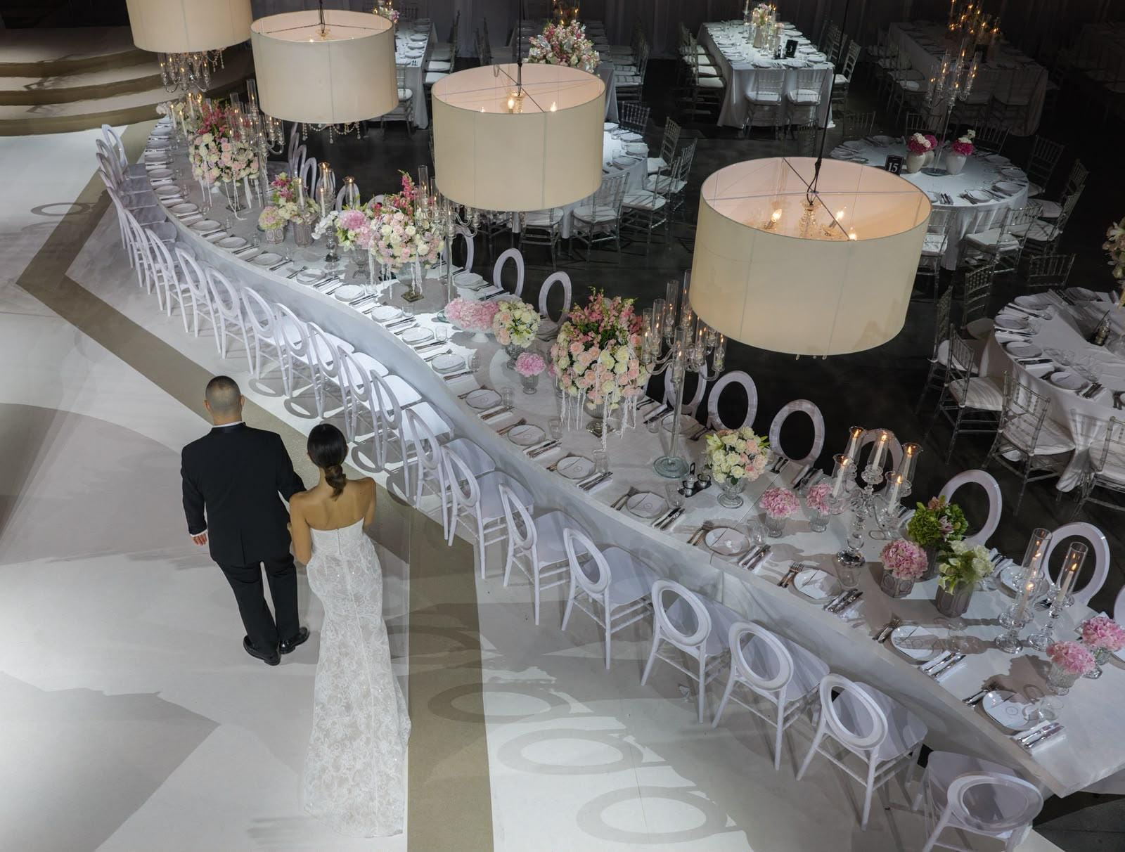 A couple in a black suit and lace gown, seen from behind, walking beside a long table under glowing drum lamps at the Hangar