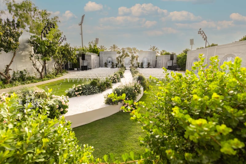 Winding path across a lawn under a blue sky with clouds, bushes in the foreground and white arched facades beyond