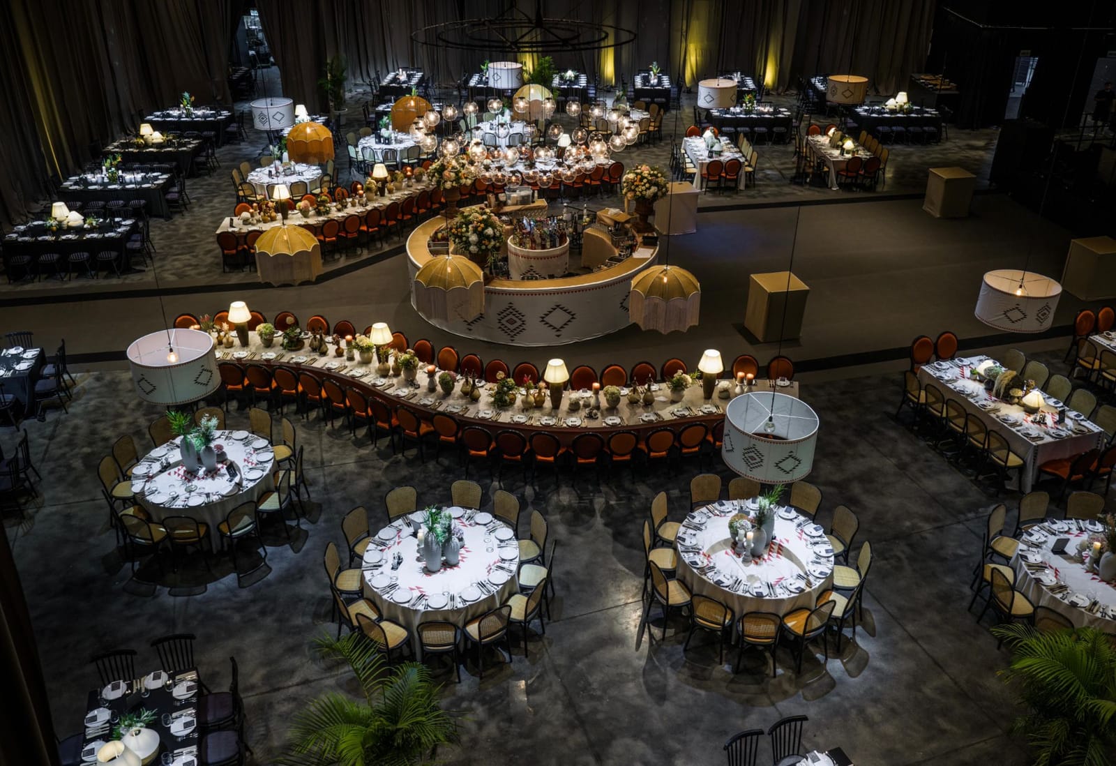 Aerial view of the Hangar set for an event: a round central bar, a winding table with orange velvet chairs, round tables and oversized lampshades