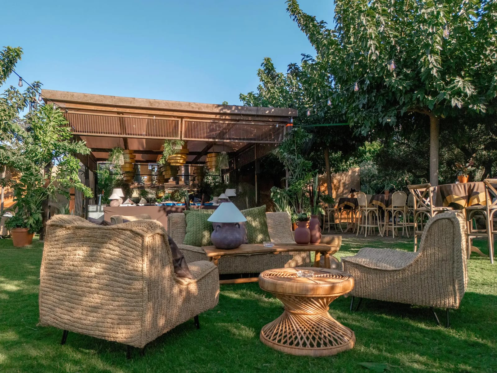 Low-angle view of a lawn lounge with wicker armchairs and a rattan table, with a wooden bar pavilion and set tables behind