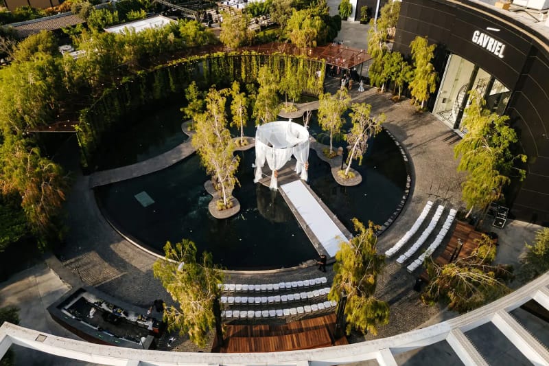Golden-hour aerial view of the round black pond at the Shulhan, with a white gazebo chuppah, white aisle and terraced seating