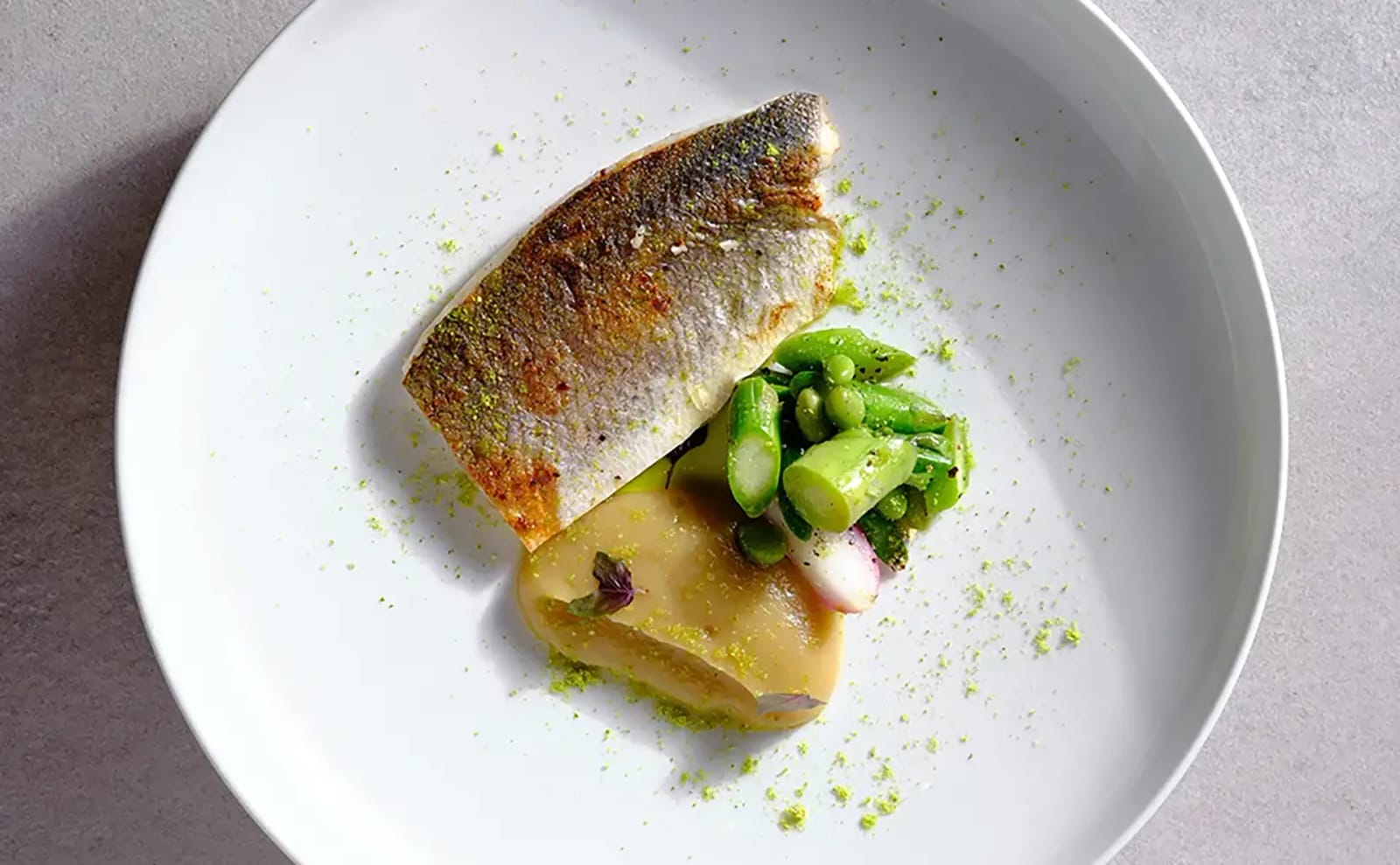 Top-down view of a crisp-skinned seared fish fillet with asparagus, pea salad, pale purée and radish on a white plate