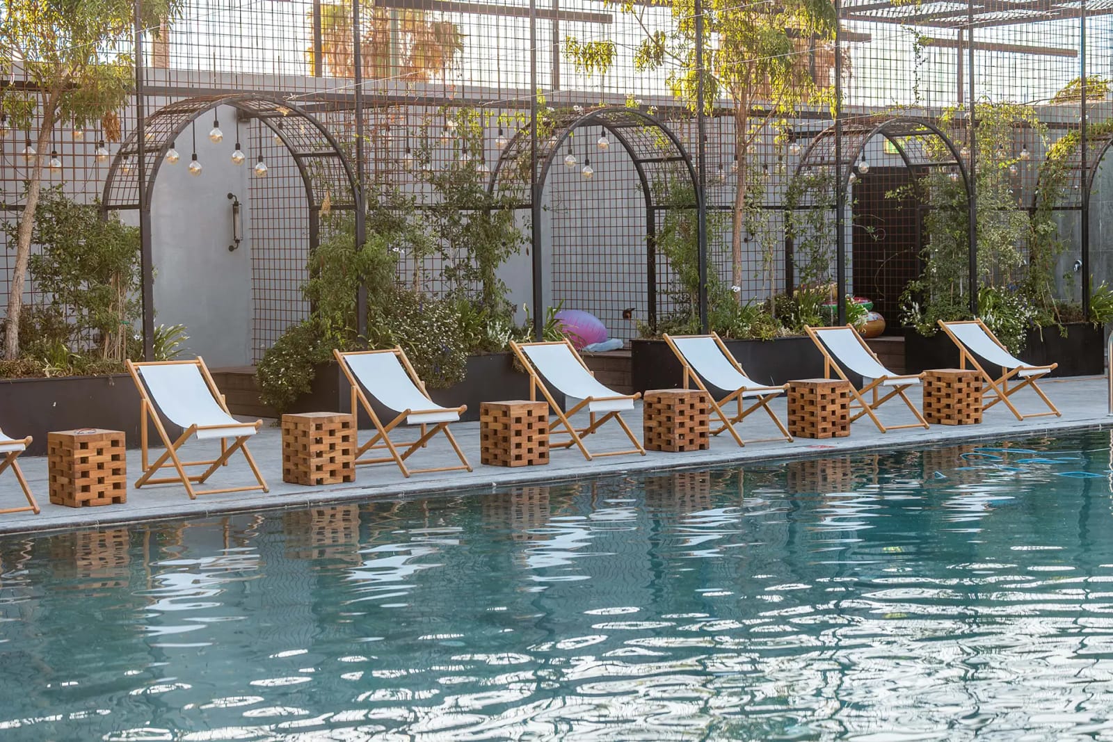 Transats blancs et tables cubes en bois au bord de la piscine du Bustan, devant des treillis d'acier noir en arche