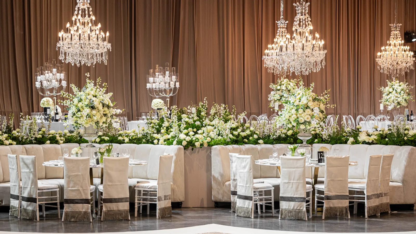 The Hangar in white and champagne, with crystal chandeliers before taupe drapes, a low white-rose garland and round tables
