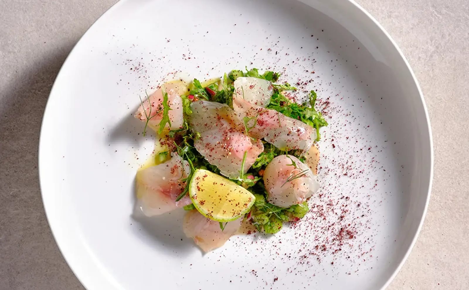 Top-down view of thinly sliced raw fish with a lime half and herb salad, dusted with dark red spice, on a white plate