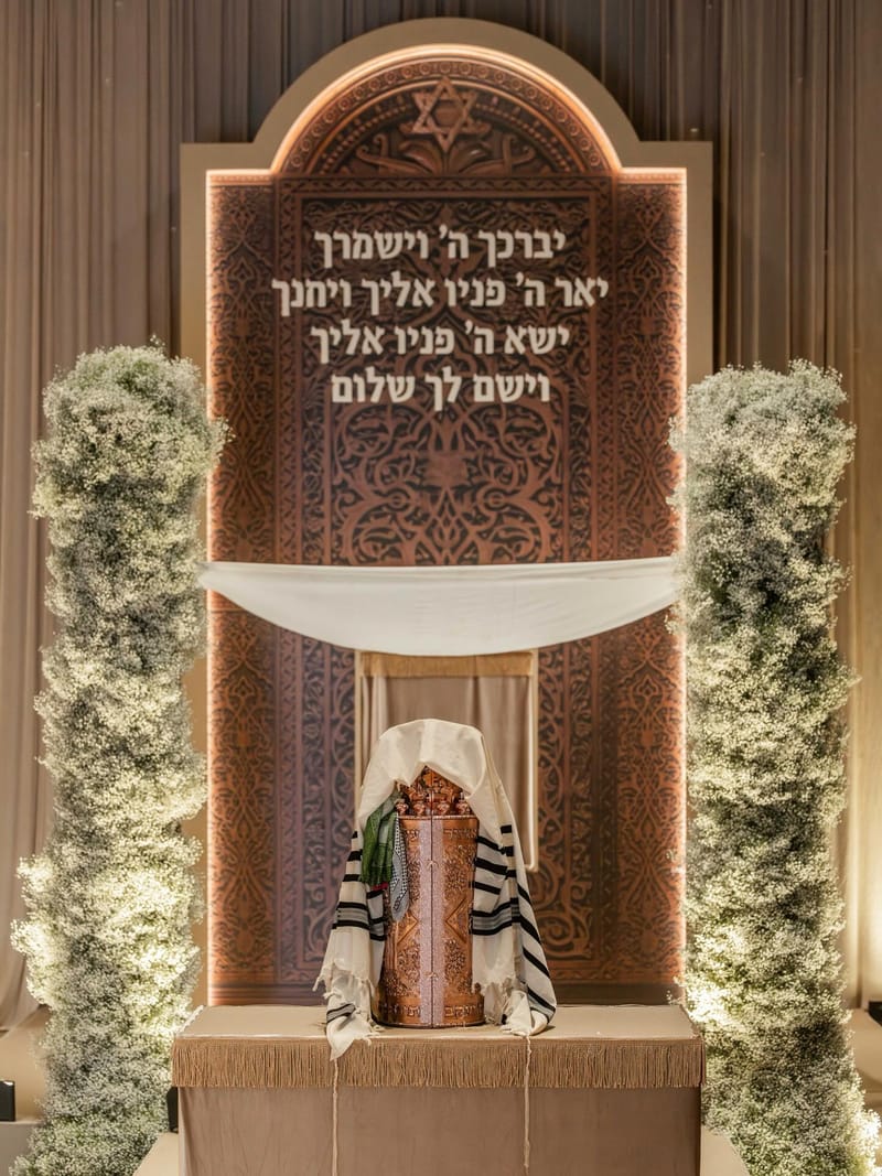 קשת עץ מגולפת עם ברכה בעברית בין שני עמודי פרחי גיפסנית גבוהים, ושולחן מכוסה בד עם ספר תורה