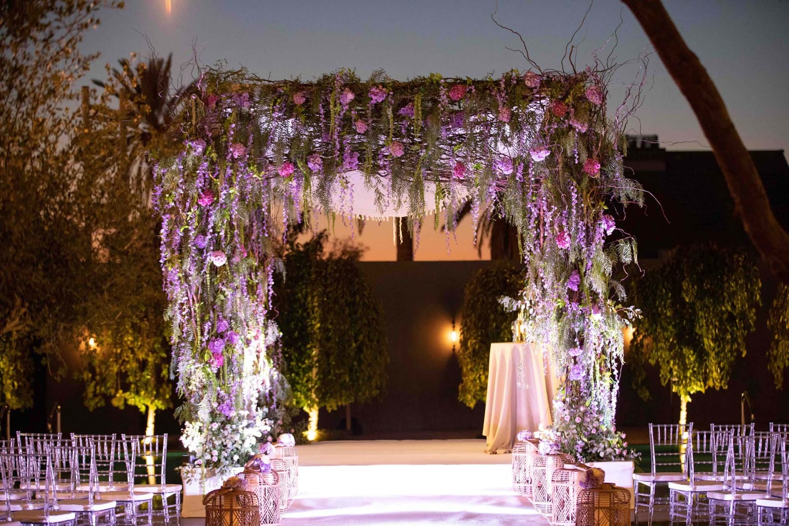 Cérémonie au crépuscule sous une houppa tressée de fleurs violettes et roses et de branches, avec bougies et chaises éclairées