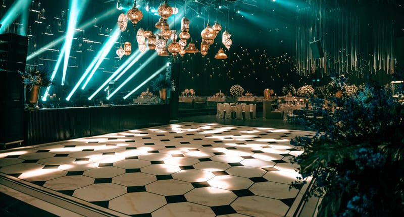 Piste de danse vide au carrelage noir et blanc, faisceaux turquoise et grappes de suspensions en cuivre, dans la salle du Shulhan