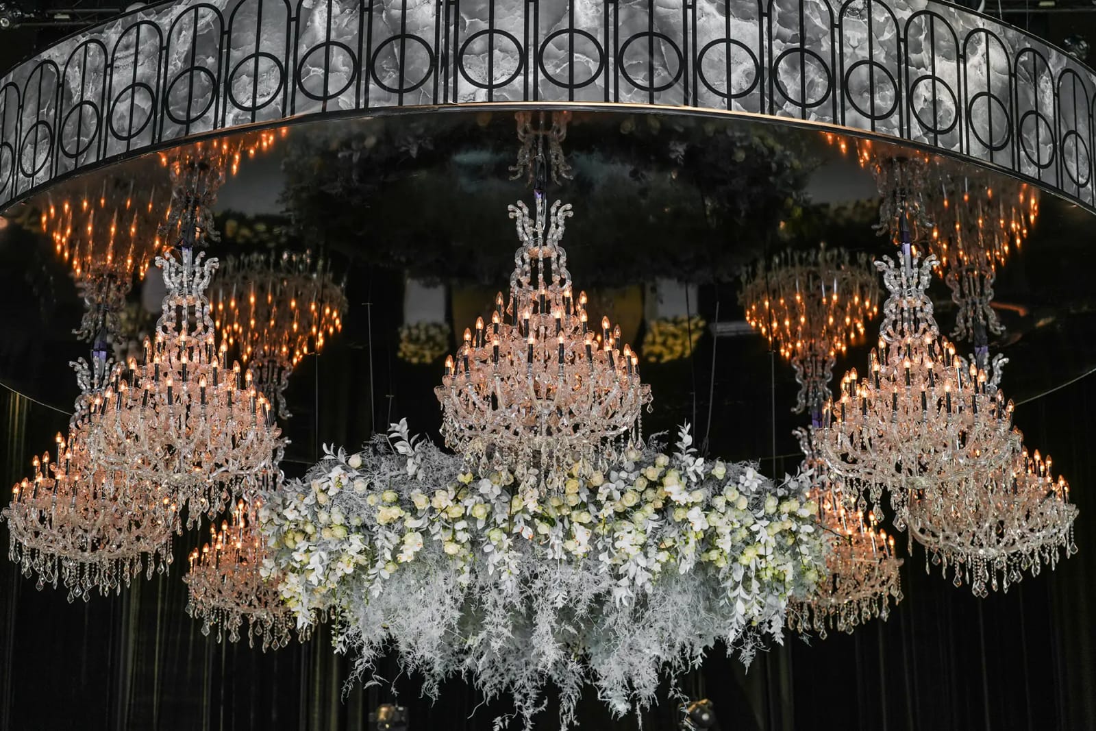 Ornate crystal chandeliers with amber bulbs beneath a mirrored truss ring, with floating white flowers and trailing greenery