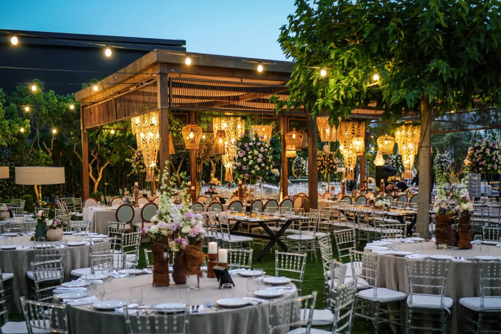 Jardin du Shulhan à l'heure bleue, pergola de lustres lumineuse derrière une table ronde fleurie et des rangées de tables dressées