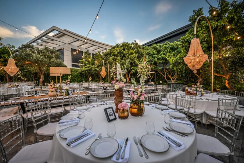 Jardin du Shulhan en fin d'après-midi, table ronde dressée, lampe-lustre en cuivre sur pied haut et longues tables sous les arbres