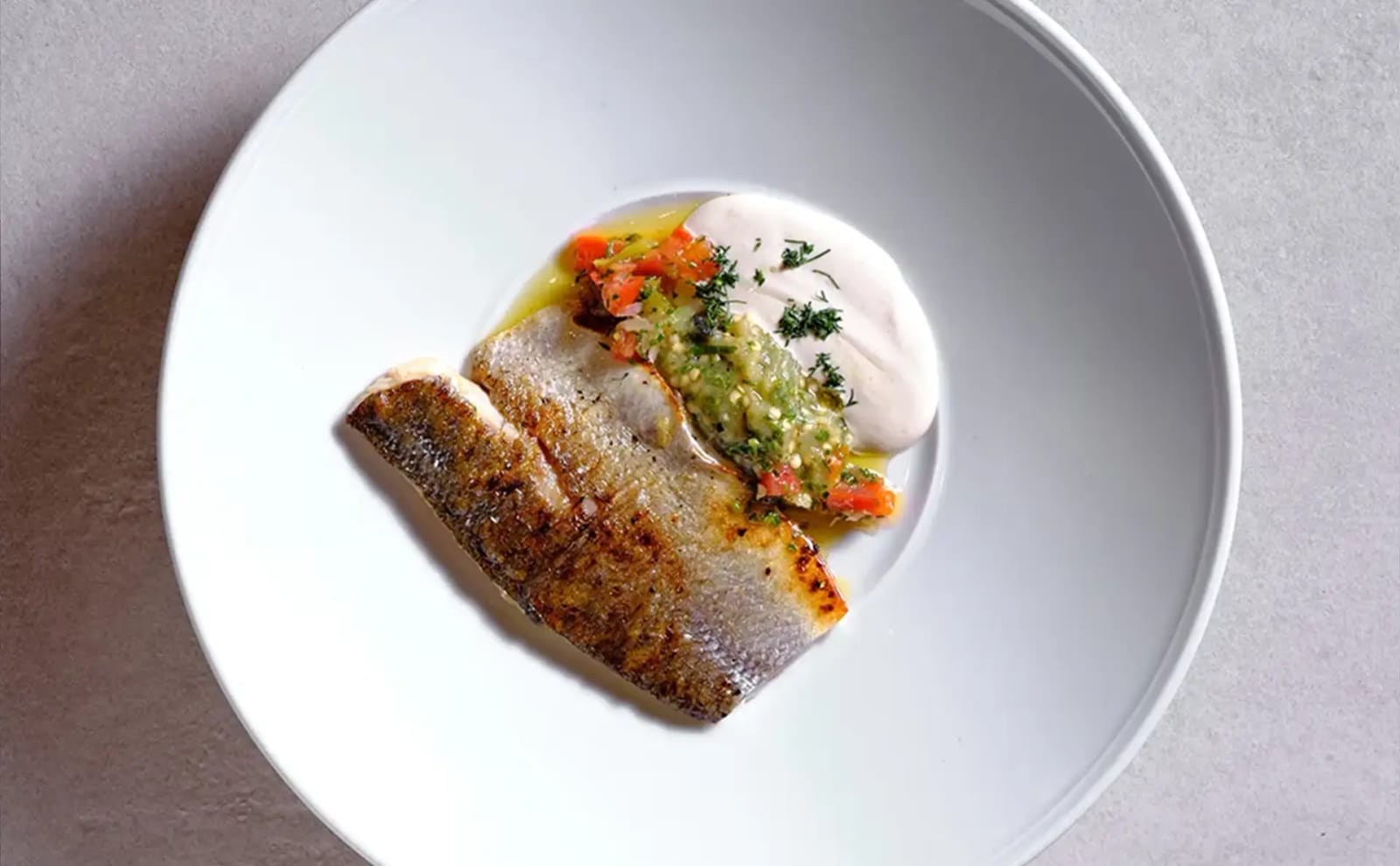 Top-down view of a crisp-skinned white-fish fillet with green and tomato relish and a dollop of cream on a white plate