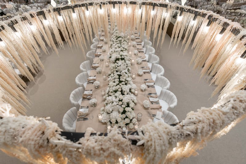 High-angle view of a long table with a floral runner at the center of a lit ring of hanging white plumes, at the Hangar
