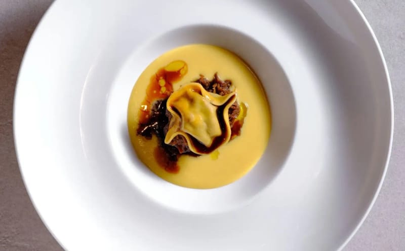 Top-down view of a filled pasta parcel with a dark braised-meat reduction and orange oil on golden cream in a white bowl