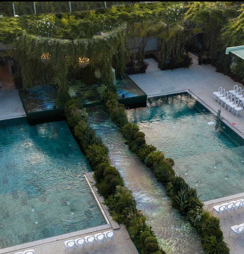 Vue en hauteur de deux piscines turquoise séparées par un canal bordé de haies, sous un auvent couvert de vigne