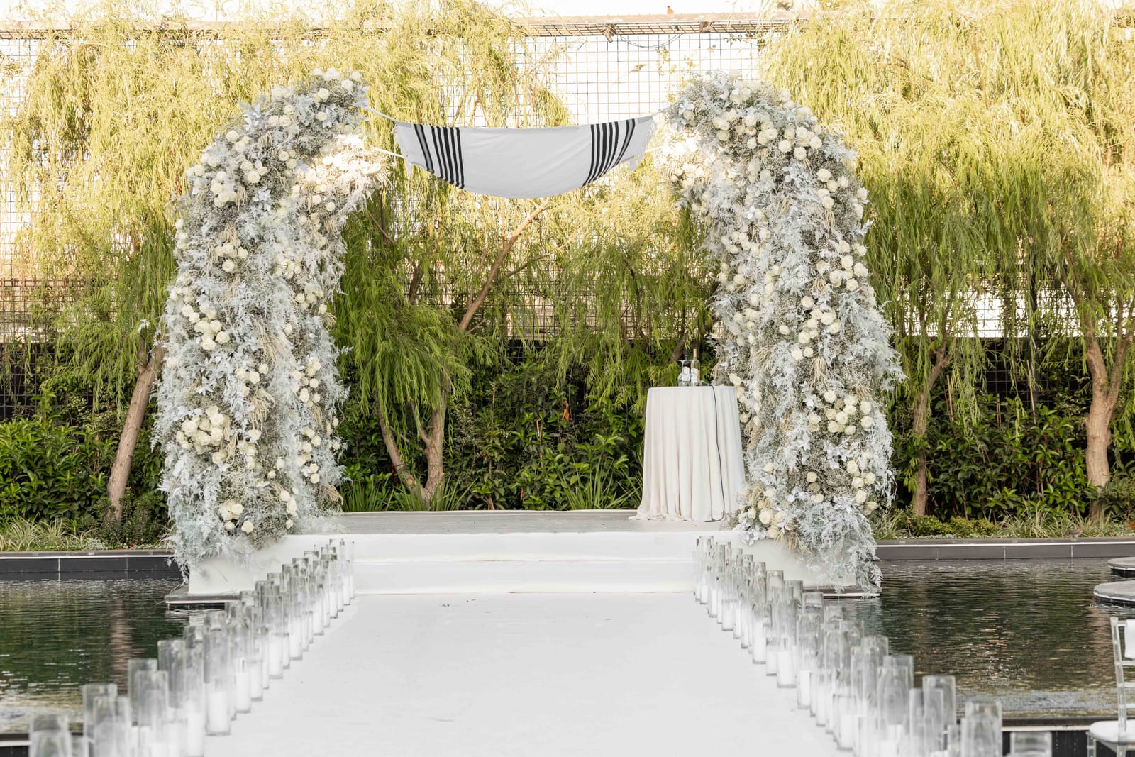 Symmetrical view down a candle-lined white aisle over dark water to a two-arch frosted floral chuppah and weeping willows