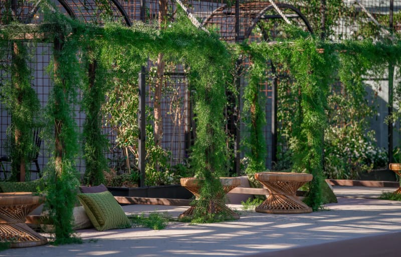 Salon de pelouse en plein jour, entre de hautes arches de treillage noires couvertes de verdure, bas tabourets en bois