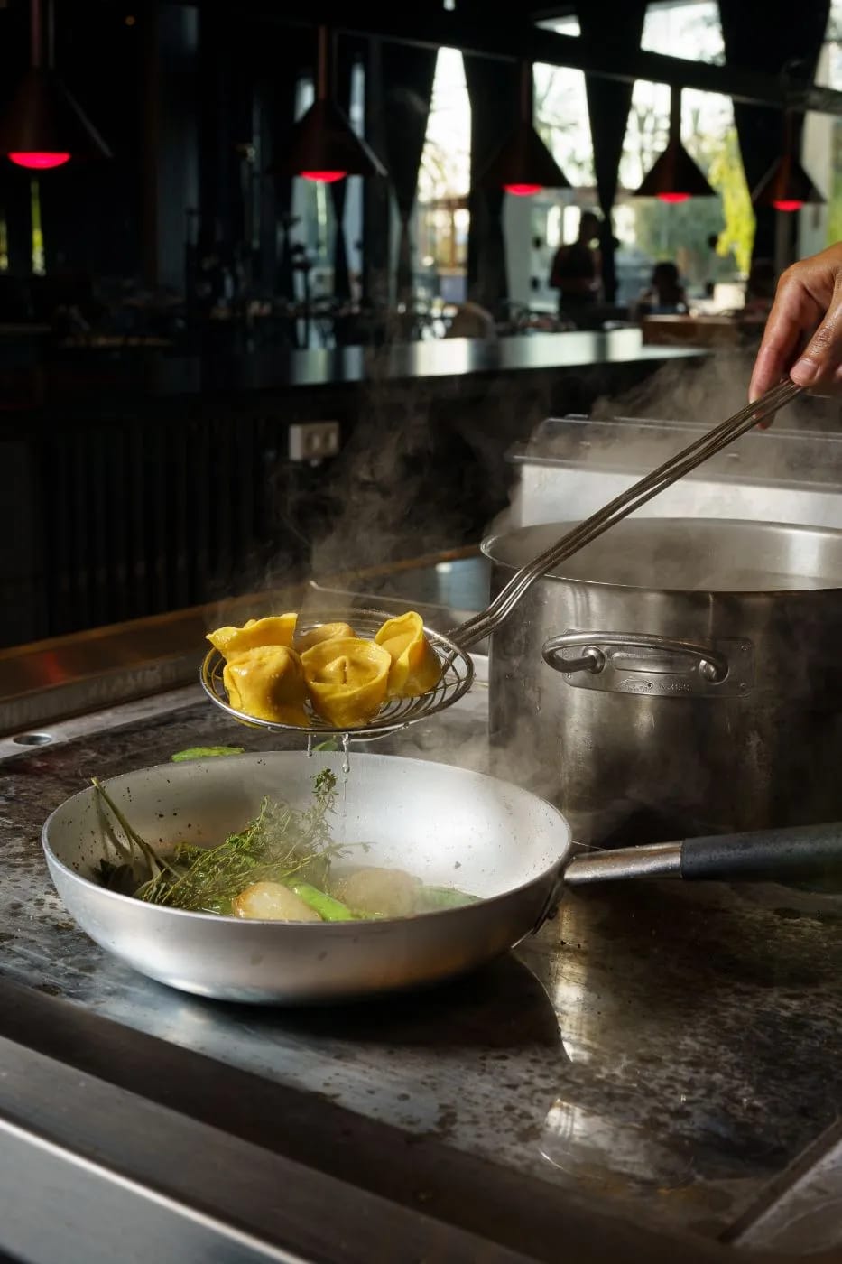 A hand lifting filled pasta on a long-handled spider strainer over a pan, with a steaming pot and red pendant lamps behind
