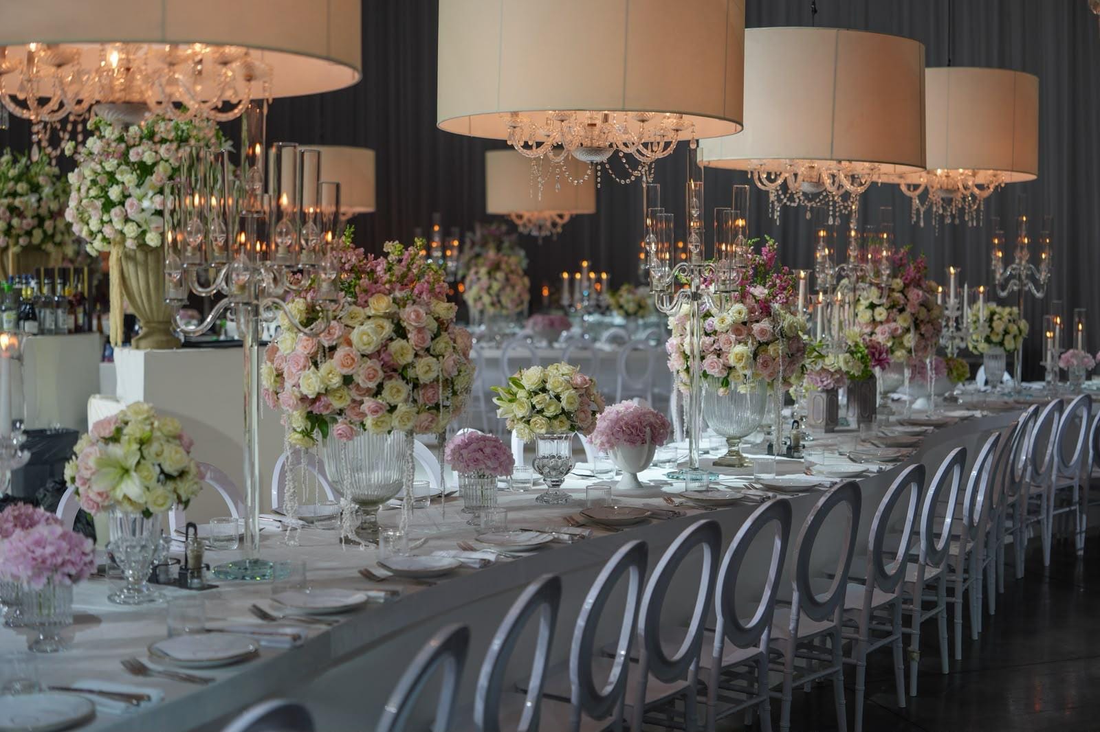Long table at the Hangar with soft pink, coral and green flowers, candelabras, white open-back chairs and glowing drum lamps