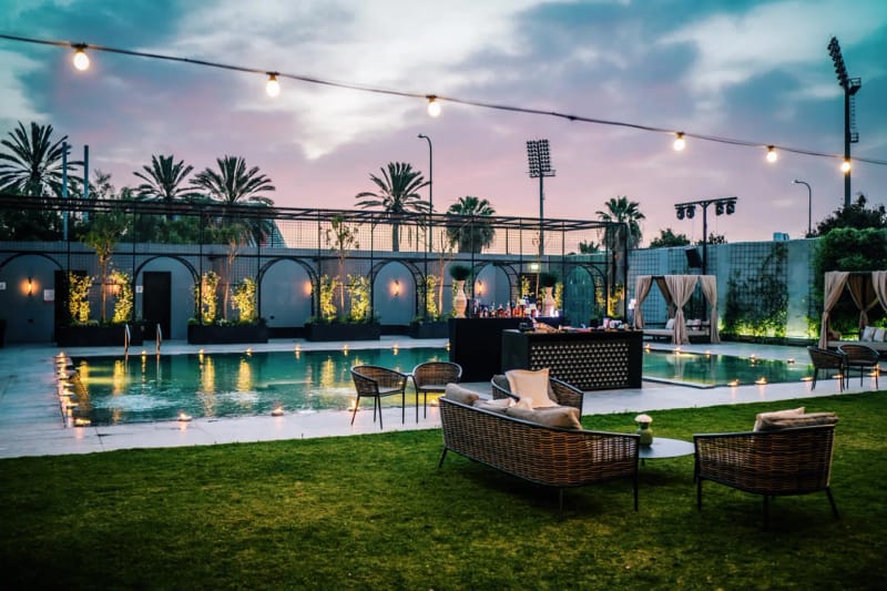 Crépuscule sur une piscine doucement éclairée, mur de jardin à arches, jardinières illuminées, mobilier en osier, guirlandes