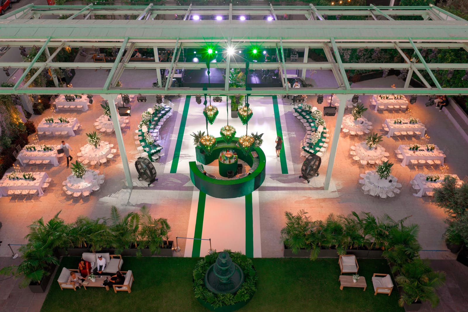Vue en hauteur le soir d'un événement de jardin sous pergola en acier, tables rondes autour d'un bar vert central, salon