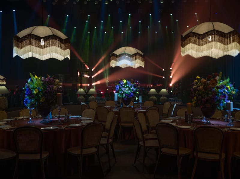 Dark Hangar hall with green, red and blue light beams behind a stage, fringed pendants and round tables with flowers