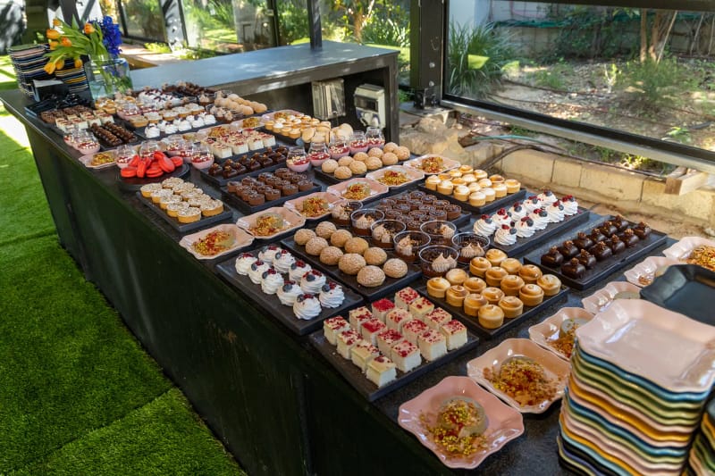 Longue table de desserts avec petits fours, tartelettes, chocolats et petits gâteaux sur plateaux sombres, devant une paroi vitrée