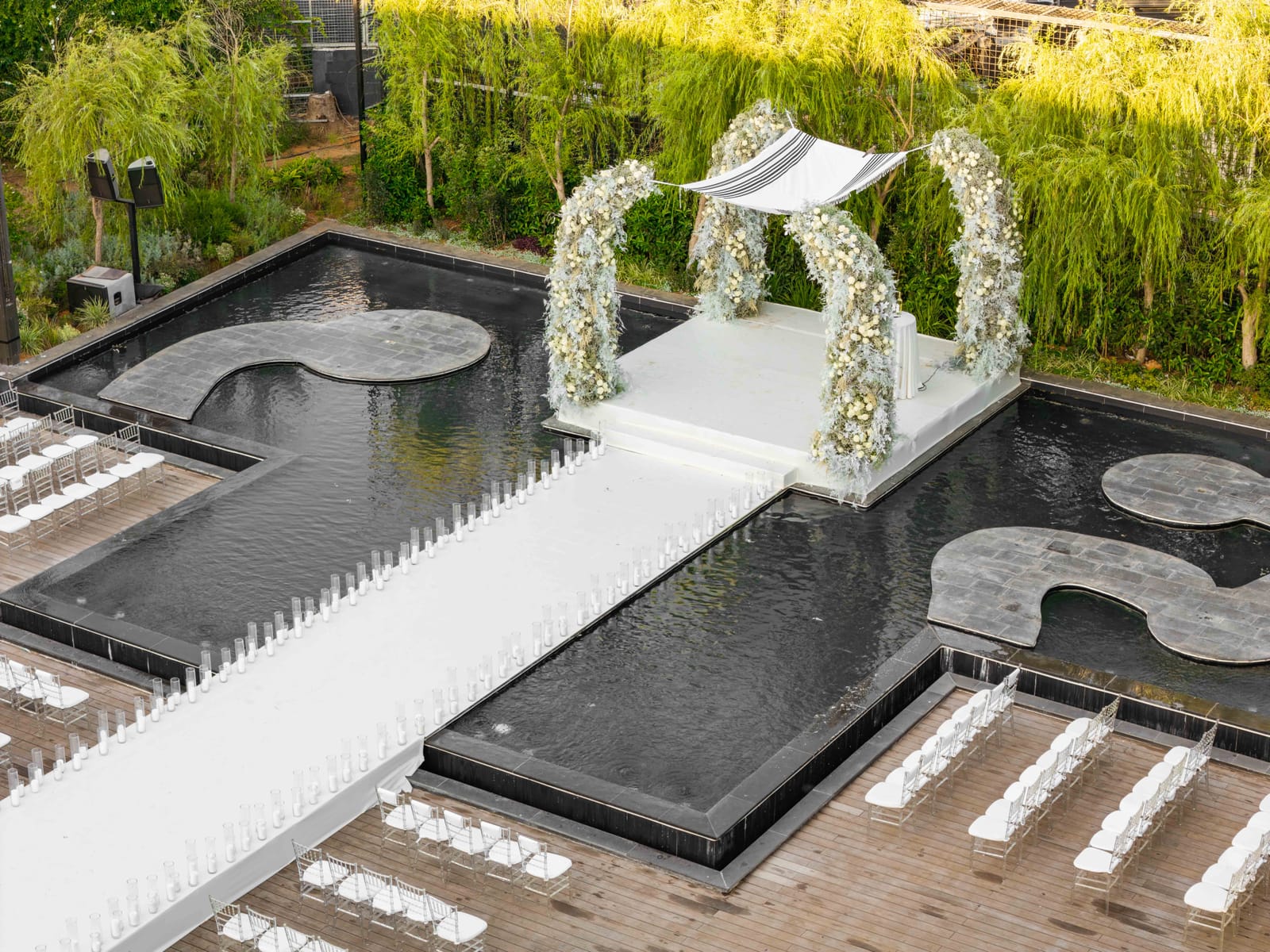 Aerial view of a floral chuppah on a white platform between two black water basins, with a candle-lined white aisle