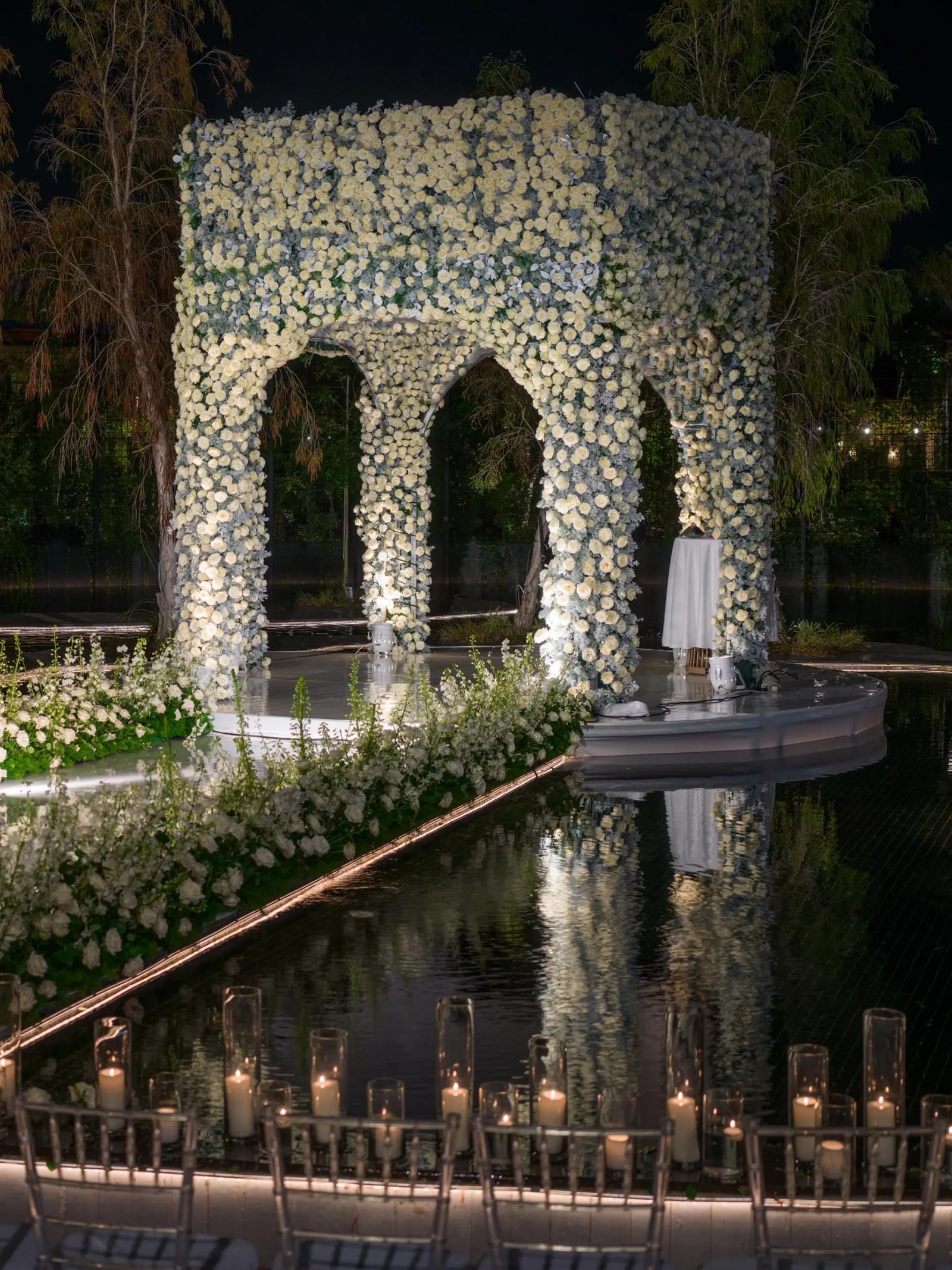 White-rose arched chuppah seen from the water's edge, with a floral walkway, candle cylinders and clear chairs at night