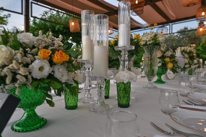 Close-up of white and orange flower arrangements in emerald glass vases, with tall candles on a set table
