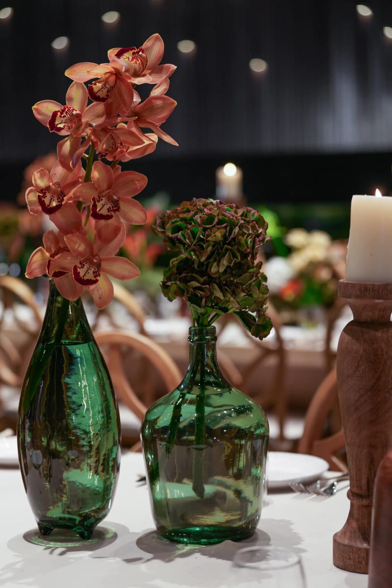 Two green glass bottles holding orange orchids and a hydrangea, beside a wooden candlestick and candle on a white table