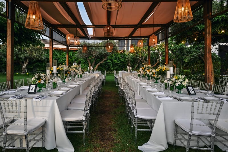 Two long rows of tables with clear chairs on either side of a grass aisle under a pergola with hanging lanterns