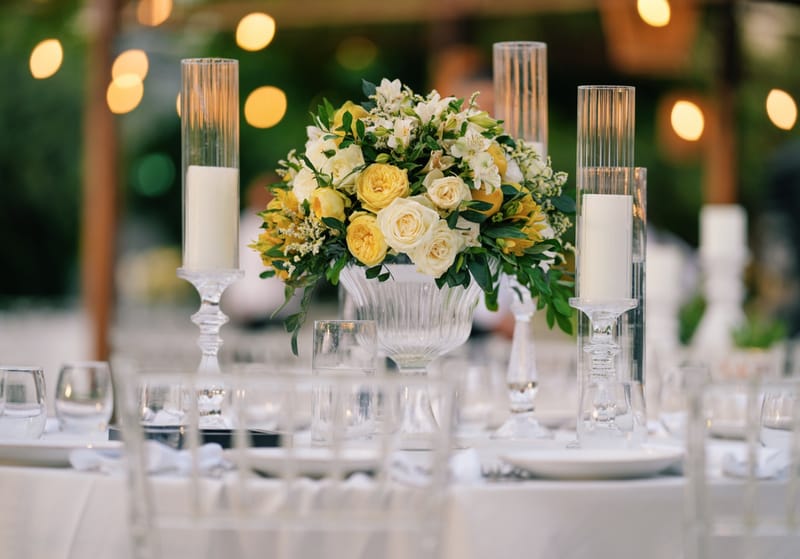 Bouquet of yellow and white roses in a glass bowl between white candles in glass cylinders, with blurred lights