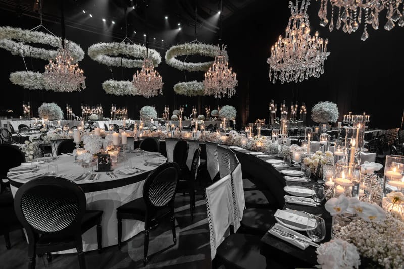 Salle sombre avec anneaux de gypsophile et lustres en cristal suspendus au-dessus de tables rondes, fleurs blanches et bougies