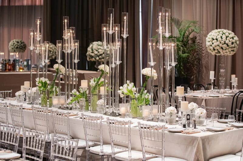 Longue table au Shulhan avec hauts photophores transparents et fleurs blanches, devant des rideaux rose pâle
