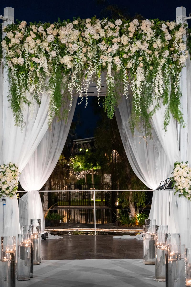 Gros plan d’une houppa au dais de fleurs blanches et de verdure retombante, voiles blancs et lanternes en verre