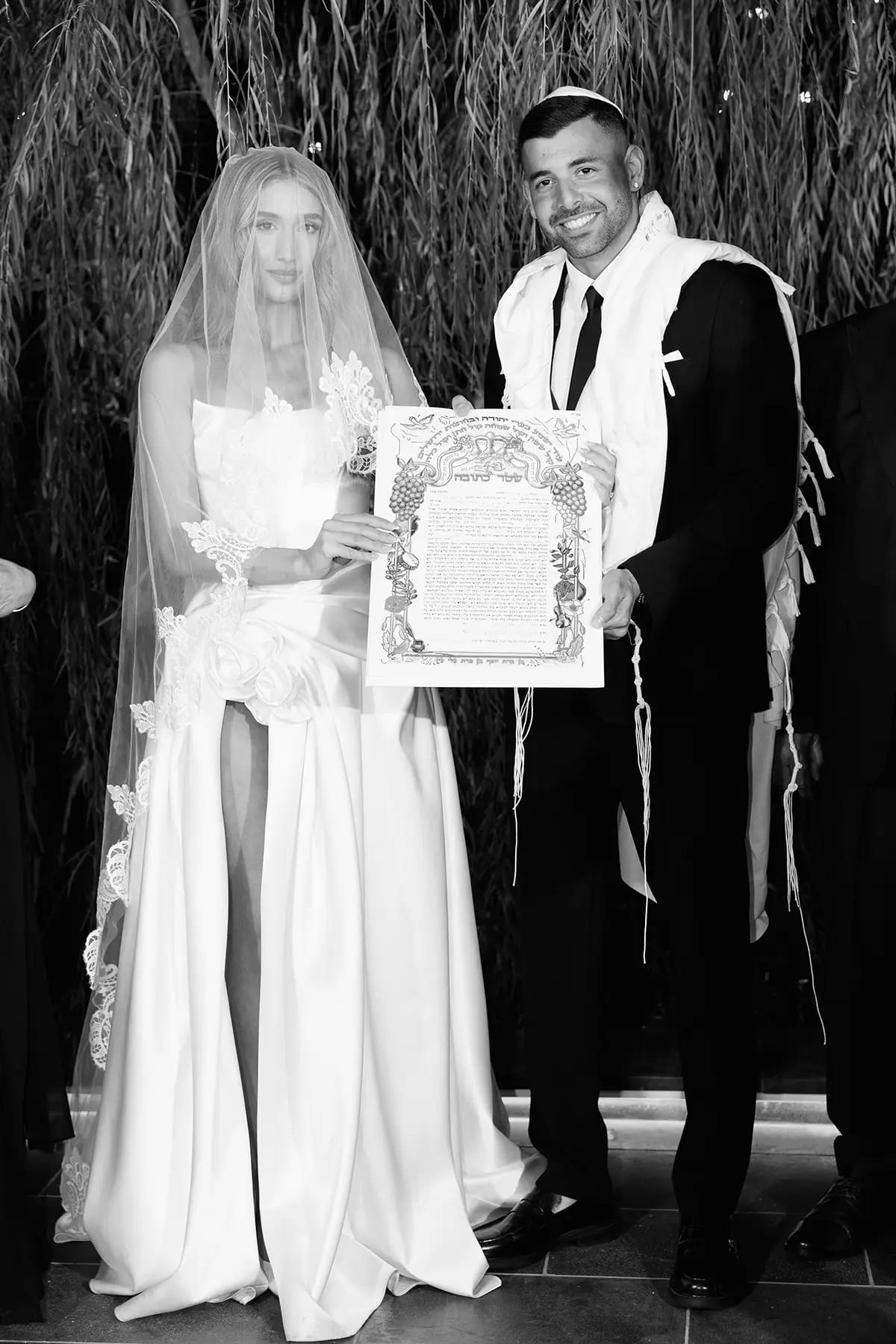 Image en noir et blanc d'une mariée voilée et d'un marié en tallit tenant une ketouba ornée, des saules derrière eux