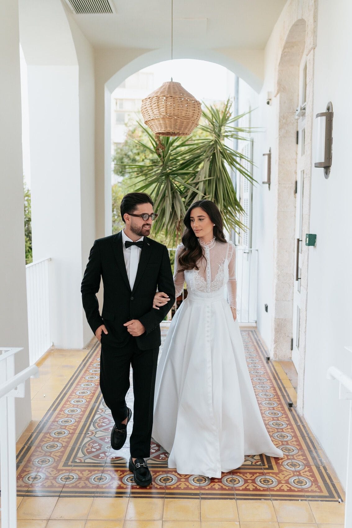 Un couple marchant dans un couloir à arcades et carreaux décoratifs, le marié en costume noir, la mariée en robe de dentelle