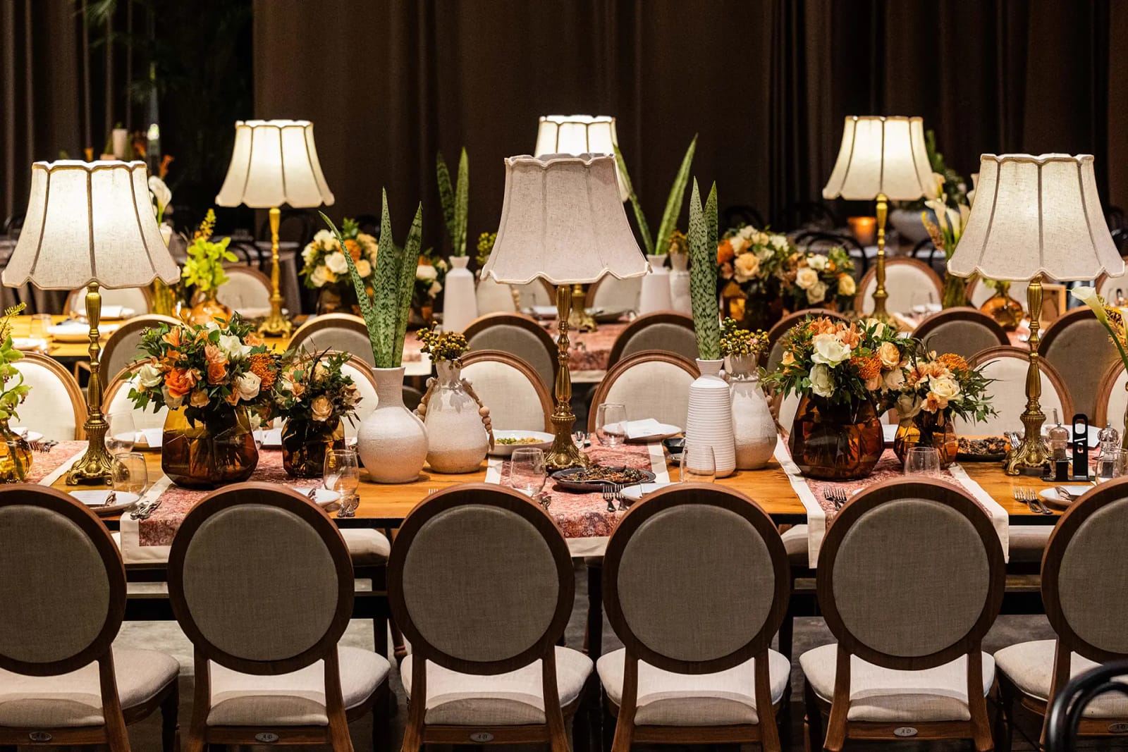 Vue de face d'une longue table avec lampes à abat-jour, fleurs orange, vases blancs et chaises à dossier rond