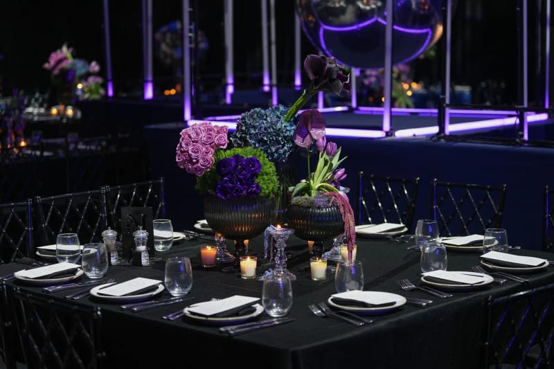Black dining table set with a purple and green floral centerpiece, with violet-lit glass frames behind