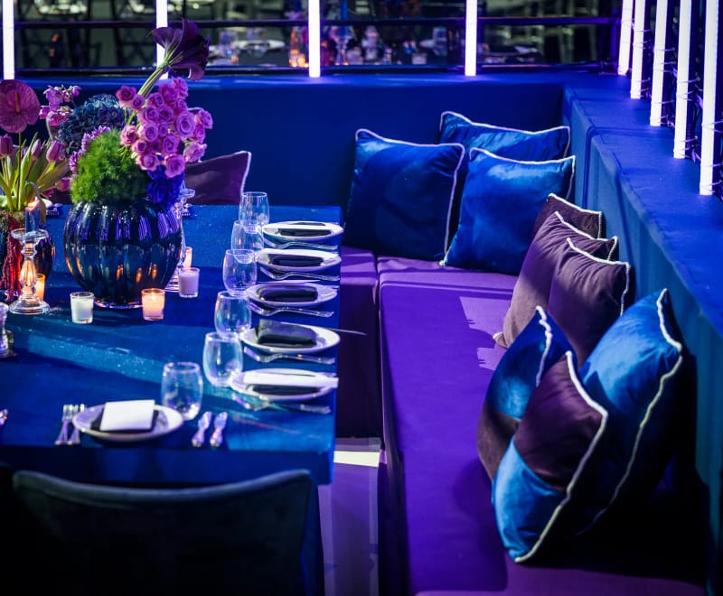 Blue velvet banquette with purple and blue cushions beside a set table with purple flowers, with neon tubes behind