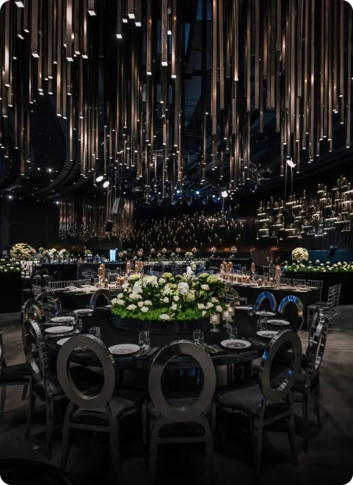 Salle sombre aux tables rondes, chaises argentées à dossier rond et fleurs blanches sous un plafond de tubes lumineux