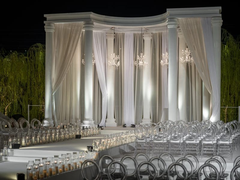 Gros plan d'un pavillon blanc à colonnade et voilages, rangées de chaises transparentes et bougies devant, de nuit