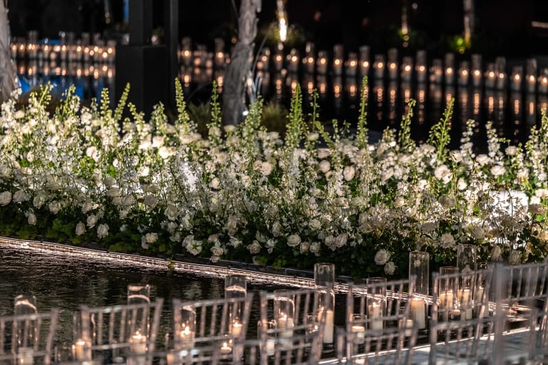 Dense bordure de fleurs blanches au-dessus d'une eau sombre avec des rangées de bougies et des chaises transparentes devant