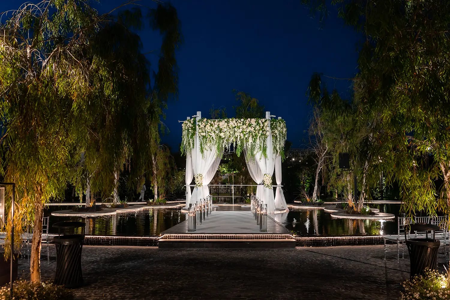 Jardin de nuit aux arbres éclairés et passerelle blanche sur l’eau menant à une houppa au loin